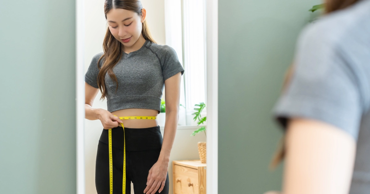 Woman measuring her waist with a yellow tape measure in front of a mirror for Weight Loss Injections in Allen, TX.